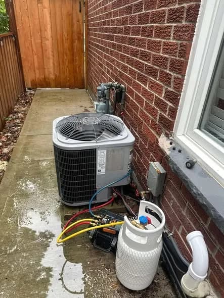 Technician's manifold gauges and a refrigerant cylinder connected to an outdoor air conditioning unit in a narrow side yard, on wet ground during rain