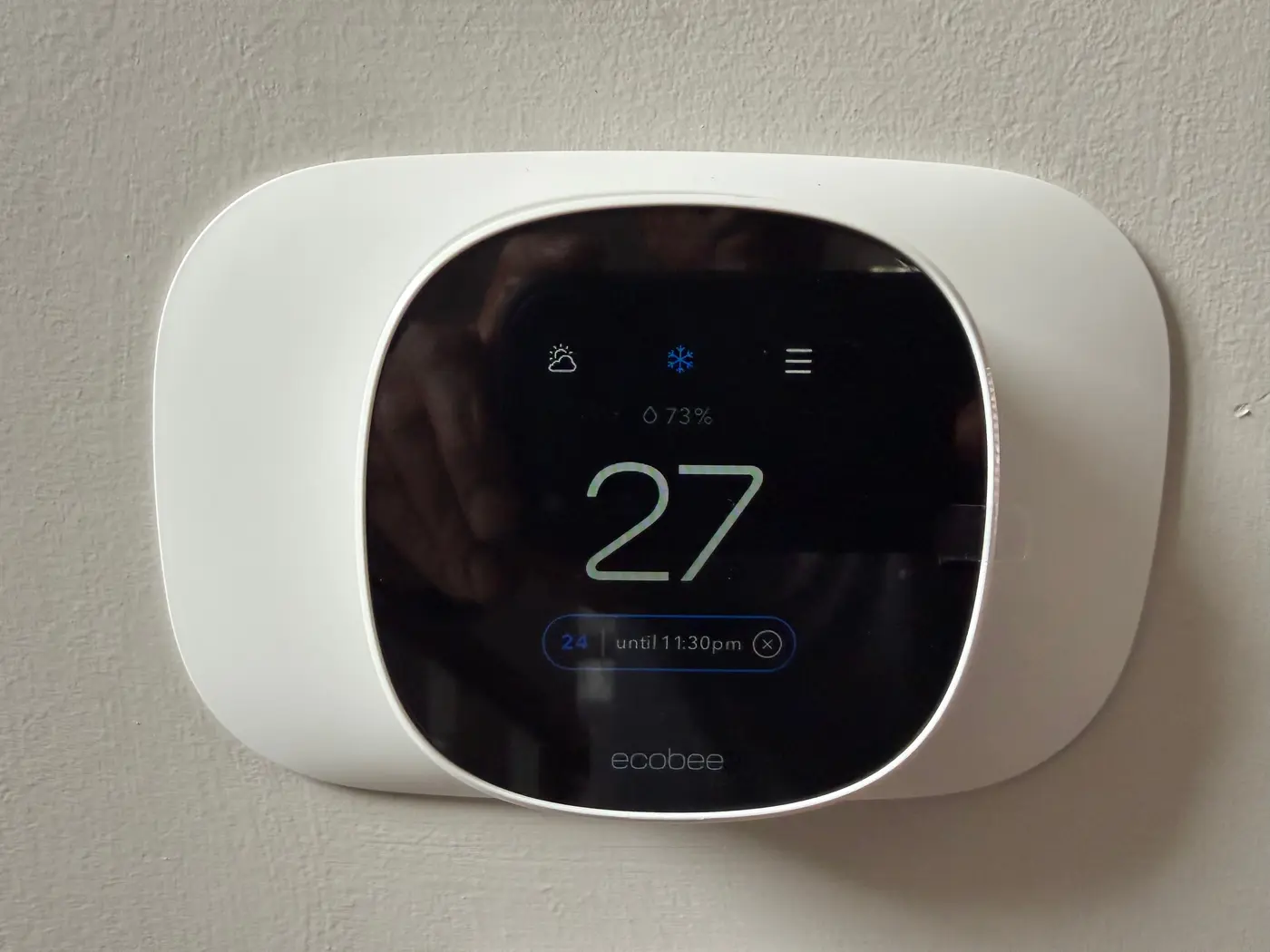 ecobee smart thermostat mounted on a wall, its display showing the room at 27 degrees Celsius, holding a 24 degree setpoint until 11:30pm