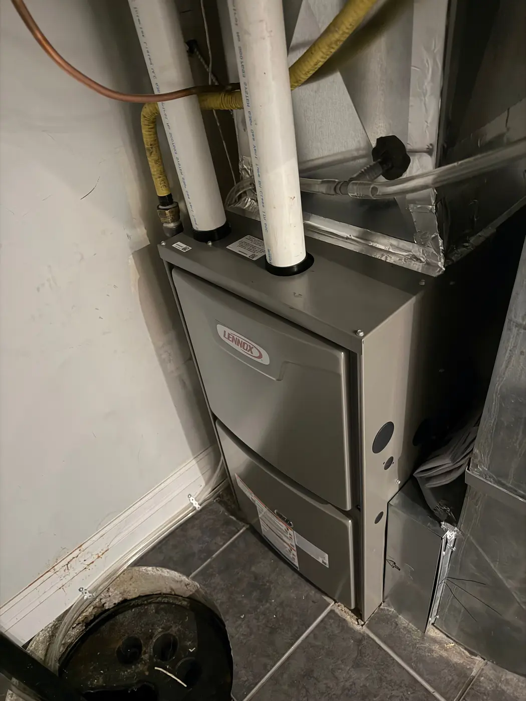 New Lennox condensing gas furnace installed in a narrow utility closet, with two white PVC vent pipes, a yellow flexible gas connector and a sealed sheet-metal plenum above it