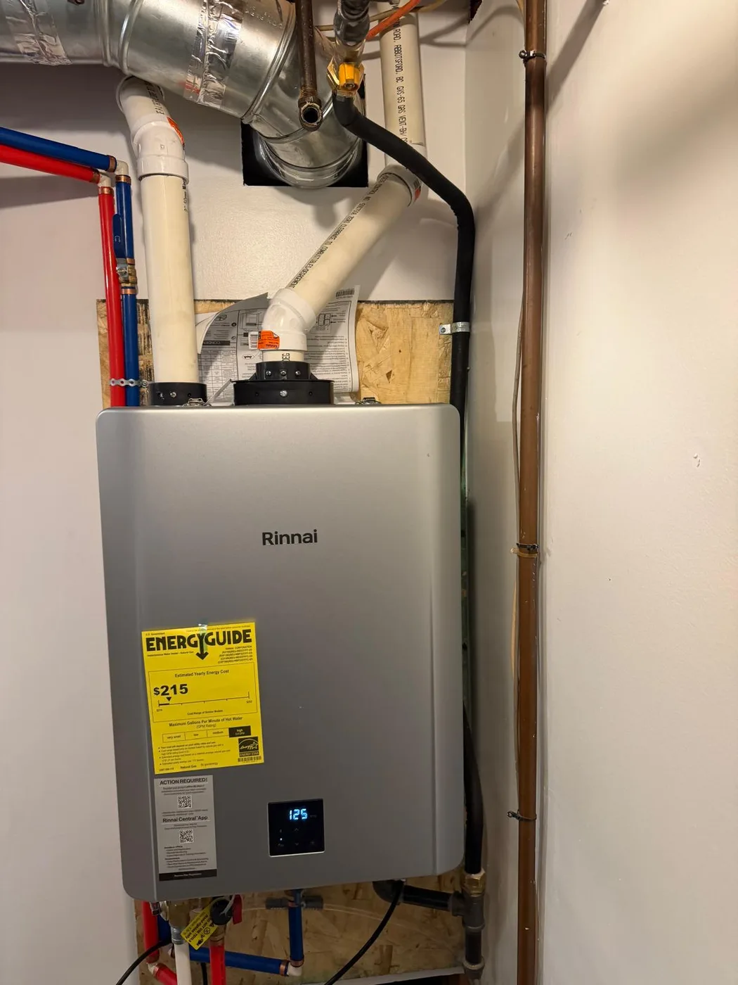 Rinnai tankless gas water heater mounted on a plywood backer board in a utility closet, with two white PVC vent pipes rising from the top and red and blue water lines to the left