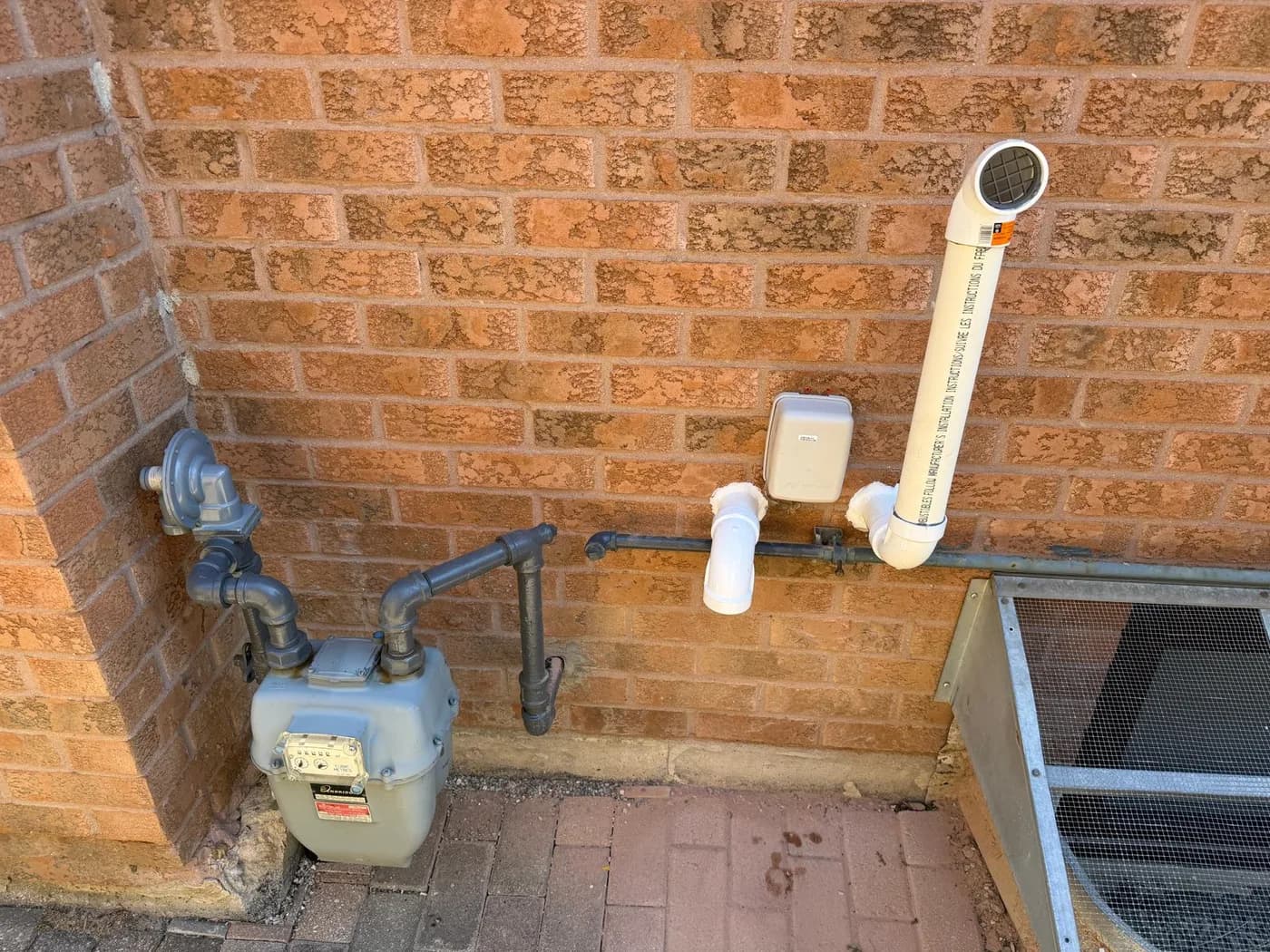 Two white PVC vent terminations coming out through a brick wall beside the gas meter, one turned down and one rising to a screened cap