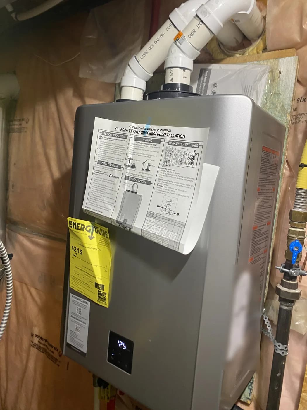 Rinnai tankless water heater mounted on an unfinished basement wall, with two white PVC vent pipes rising from the top and the installation sheet still taped to the front
